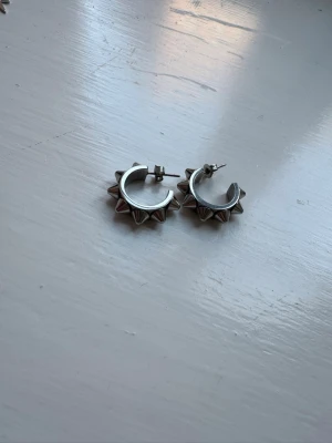 Silvriga örhängen med nitar - Säljer ett par coola silvriga örhängen i metall med rund form och spetsiga nitar runt hela ringen. De har en edgy och unik design som verkligen sticker ut. Perfekta för dig som gillar att addera lite attityd till din stil.