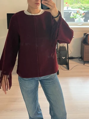 Burgundy tröja med fransar - Säljer en vinröd tröja med vida ärmar och långa fransar vid ärmsluten. Tröjan har en rak passform och rund halsringning. Perfekt för dig som vill ha en unik och trendig look.