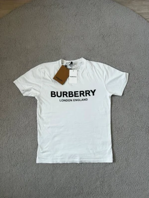 Vit Burberry t-shirt med logga - Snygg vit t-shirt från Burberry med stor svart logga och texten 'London England' på bröstet. Klassisk rund halsringning och korta ärmar. Tillverkad i mjuk bomull för en bekväm känsla. Perfekt för dig som gillar stilrena och exklusiva plagg.Fraktar samma dag eller nästa📦🚚 Aktiv hela tiden✅ hör av dig om du undrar något eller vill ha fler bilder⁉️