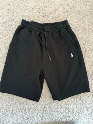 Svarta shorts från Polo Ralph Lauren - Svarta shorts från Polo Ralph Lauren med elastisk midja och snörning. Klassisk logga broderad i vitt på vänster ben. Mjukt material och två sidofickor gör dem perfekta för chill dagar. Enkel och stilren design som funkar till det mesta.