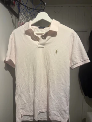 Ljusrosa pikétröja från Polo Ralph Lauren - Säljer en ljusrosa pikétröja från Polo Ralph Lauren med klassisk krage och knappslå. Tröjan har det ikoniska broderade logotypen på bröstet och är i slim fit-modell. Perfekt för dig som gillar stilrena och tidlösa plagg.
