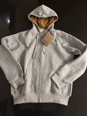  Burberry Zip-hoodie  - Står xxl passar s/m alltså inte vad som står på taggen, Snygg grå hoodie med dragkedja från Bur. Klassisk huva med snörning och innerfoder i det ikoniska Bur-mönstret. Broderad logga på bröstet och två fickor framtill. Mjuk bomullsblandning och ribbade muddar för extra komfort.
