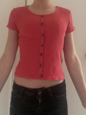 Ribbad rosa topp från H&M - Kortärmad ribbad topp i rosa från H&M med knappar framtill. Modellen är croppad och har en rund halsringning. Perfekt för en trendig och avslappnad stil.
