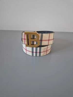  Burberry - Stilrent bälte från Burberry med klassiskt rutmönster i beige, svart och rött. Bältet har ett stort guldfärgat spänne med 'TB'-logga och är tillverkat i skinn. Perfekt accessoar för att lyfta din outfit.