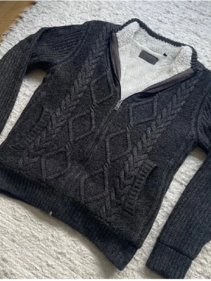 Mörkgrå stickad ziptröja med foder - Mysig mörkgrå stickad tröja med dragkedja och vitt teddyfoder på insidan. Tröjan har ett snyggt kabelstickat mönster framtill, ribbade muddar och hög krage. Perfekt för kalla dagar och riktigt skön att ha på sig. 