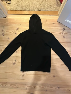 Svart 100% merinohoodie med huva - En enkel och stilren svart merinohoodie med huva och långa ärmar. Hoodien har en klassisk passform utan synliga tryck eller detaljer. Hör av dig vid minsta fundering. 