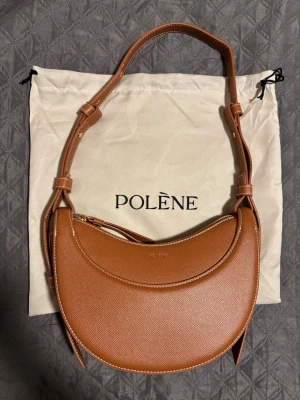 Polène brun axelremsväska i läder - Snygg brun handväska från Polène i halvmåneformad design. Väskan är tillverkad i strukturerat läder med justerbar axelrem och guldfärgad dragkedja. Perfekt för dig som vill ha en stilren och trendig accessoar.