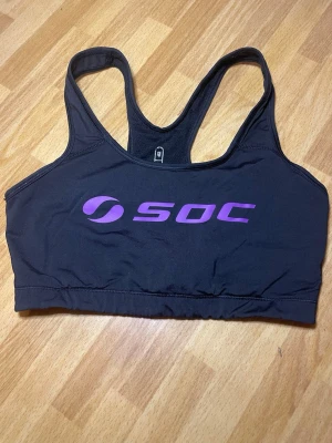Svart sport-bh från SOC - Sport-bh i svart med lila SOC-logga framtill. Bh:n har brottarrygg och bred resår under bysten för extra stöd. Tillverkad i ett stretchigt funktionsmaterial som andas, perfekt för träning och aktivitet.