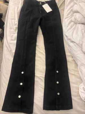 Svarta bootcut jeans med knappar - Säljer ett par svarta bootcut jeans från Cropped Flare med dekorativa silverknappar längs benen framtill. Jeansen har hög midja, utsvängda ben och klassiska fickor bak. Snygg detalj med broderad logga på bakfickan. Perfekta för en trendig look. Lappen är inte kvar 