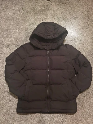 Svart pufferjacka från Jack & Jones - Säljer en svart pufferjacka från Jack & Jones med huva och dragkedja framtill. Jackan har två sidofickor och en klassisk quiltad design som håller värmen. Perfekt för kalla dagar och enkel att matcha med olika outfits.