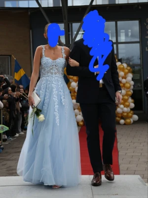Ljusblå långklänning med spetsdetaljer - Elegant ljusblå långklänning med tunna axelband och vacker spets upptill. Klänningen har en figurnära överdel med broderade blommor och en luftig kjol i flera lager. Perfekt för bal eller student. Visst slitage nedtill då den släpar lite men syns mest undertill , går att lägga upp om man är kortare än tjejen som är 178 cm. Nypris 5500 kr