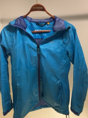 Blå vindjacka från Everest med huva - Snygg blå vindjacka från Everest med dragkedja framtill, två fickor med dragkedja och meshfoder i huvan. Jackan har en sportig look och är perfekt för blåsiga dagar. Lätt material och justerbar huva för extra komfort.