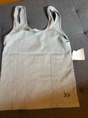 Ljusgrå linnetopp med struktur - Säljer en ljusgrå linnetopp med diskret strukturmönster och breda axelband. Toppen har en enkel och stilren design med liten logga nertill. Perfekt för en avslappnad och sportig look.
