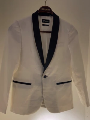 Vit kavaj med svart krage Opposuits - Stilren vit kavaj från Opposuits med svart sjalkrage och svarta detaljer vid fickorna. Enkelknäppt modell med klassisk passform och lång ärm. Perfekt för dig som vill sticka ut på festen eller middagen.