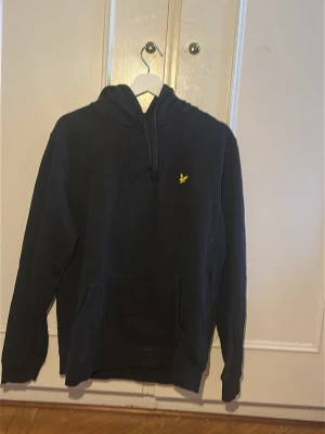 Svart hoodie från Lyle & Scott - Säljer en svart hoodie från Lyle & Scott med klassisk gul logga på bröstet. Hoodien har huva med dragsko och en stor magficka framtill. Tillverkad i mjukt bomullsmaterial och har ribbade muddar vid ärmslut och nederkant.