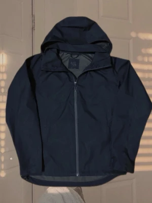 Mörkblå vindjacka med huva - Stilren mörkblå vindjacka med hel dragkedja och huva. Jackan har en minimalistisk design, långa ärmar och är tillverkad i ett lätt och vindtätt material. Perfekt för blåsiga dagar och enkel att matcha med olika outfits.