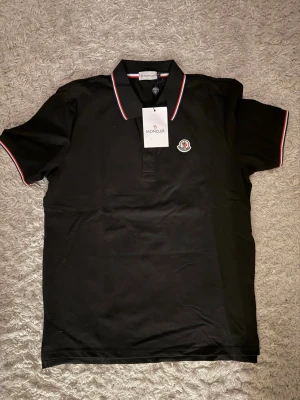 Svart Moncler piké med röd/vit kant - Snygg svart pikétröja från Moncler med klassisk krage och röd/vit kantband. Tröjan har korta ärmar och Moncler-logga broderad på bröstet. Perfekt för dig som vill ha en stilren och exklusiv look. Helt ny, absolut inte använd! Har du frågor och funderingar så är de bara o slå ett meddelande.📣 