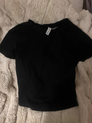 Svart croppad topp från Divided - En enkel svart croppad topp från Divided. T-shirten har korta ärmar och rund halsringning. Perfekt att matcha med höga jeans eller kjol för en clean och stilren look.