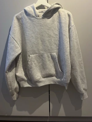 Ljusgrå hoodie från 157 - En stilren ljusgrå hoodie från 157 med klassisk huva och stor magficka. Tillverkad i mjukt sweatshirtmaterial som känns skönt mot huden. Perfekt för dig som gillar en enkel och avslappnad look. Ribbade muddar vid ärmar och nederkant för extra komfort.
