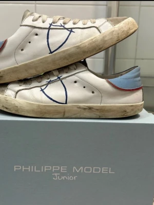 Philippe Model vita sneakers med blå detalj - Snygga vita sneakers från Philippe Model Junior med blå och röda detaljer på hälen och blått broderat märke på sidan. Skorna har klassisk låg profil, vita skosnören och är tillverkade i läder med mockadetaljer på plösen.