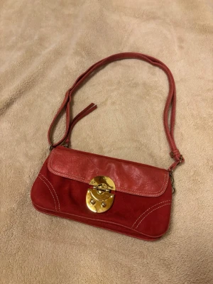 Röd axelväska med gulddetalj - Snygg röd axelväska i en mix av mocka och läder med dekorativa vita sömmar. Väskan har ett justerbart axelband. Bra använt skick, guldiga låset har några repor. Köpt i Japan 