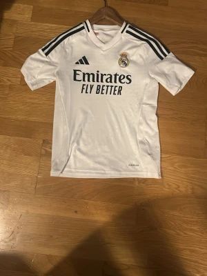 Real Madrid matchtröja  - Snygg vit Real Madrid matchtröja från Adidas med svarta detaljer och klubbmärke på bröstet. Tillverkad i lätt och ventilerande material, perfekt för fotbollsträning eller match. Hör av vid funderingar ✌️