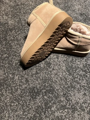 Beige fluffiga tofflor i box - Mysiga beige tofflor med mjukt foder och ovandel i mockaliknande material. De har en enkel slip-in design och ser riktigt varma ut, perfekta för att hålla fötterna sköna hemma. Kommer i originalkartong.