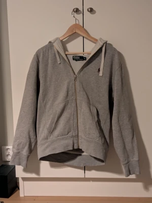 Grå zip hoodie från Polo Ralph Lauren - Klassisk grå hoodie med dragkedja från Polo Ralph Lauren. Tröjan har huva med vita snören, två fickor fram och den ikoniska lilla broderade loggan på bröstet. Tillverkad i mjukt bomullsmaterial som är perfekt för chill dagar.