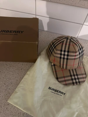 Burberry klassisk rutig keps - Säljer en ikonisk Burberry keps i klassiskt beige rutigt mönster med svarta, vita och röda detaljer. Kepsen har justerbar rem bak och Burberry-logga. Tillverkad i bomull och levereras med originalpåse och box. Perfekt för dig som vill ha en stilren och trendig accessoar.