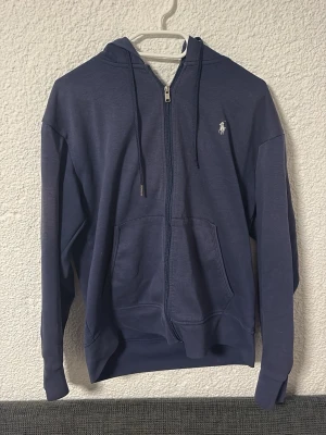 Marinblå hoodie från Polo Ralph Lauren - Snygg marinblå hoodie från Polo Ralph Lauren med dragkedja framtill, justerbar huva och fickor.  Defekter ser ni på första o sista bilden. Ena metall saken från ena snöret är borta och sen den man hänger upp tröjan med är av. Därav det billiga priset!