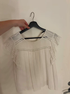 Vit blus med spetsdetaljer - Söt vit blus med rund halsringning och korta ärmar i broderad spets. Blusen har dekorativa hålbroder soppar och volangdetaljer längs axlarna. Luftigt material som ger en romantisk vibe, perfekt till jeans eller kjol. Vet ej märket, köpt på vintage butik