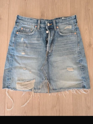 Blå jeanskjol med slitningar - Säljer en blå jeanskjol från Levi's med rå kant och slitna detaljer framtill och baktill. Kjolen har klassiska fem fickor, knappgylf och en kort passform. Perfekt för dig som gillar en avslappnad och trendig stil.