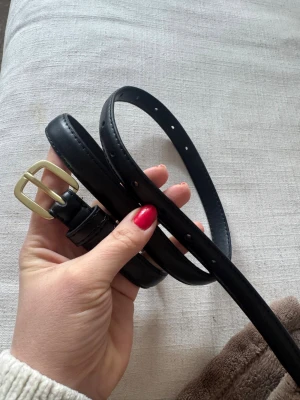 Svart smalt bälte med guldfärgat spänne - Stilrent svart bälte i smal modell med klassiskt guldfärgat spänne. Perfekt accessoar för att lyfta din outfit. Bältet har flera hål för justerbar passform och är gjort i ett slätt material.