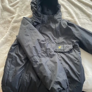 Svart vindjacka från Lyle & Scott - Snygg svart vindjacka från Lyle & Scott med huva och dragkedja framtill. Jackan har en stor ficka på magen med gul logga, samt elastiska muddar vid ärmslut. Perfekt för blåsiga dagar och enkel att matcha med olika outfits.