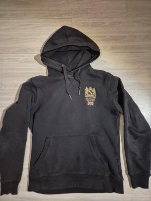 Svart hoodie från Morris Stockholm - Snygg svart hoodie från Morris Stockholm med guldfärgat broderat emblem och brittisk flagga på bröstet. Klassisk känguruficka framtill och justerbar huva med snören. Perfekt för en avslappnad och stilren look.