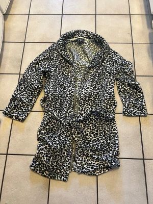 Leopardmönstrad jacka storlek XL/XXL - Mysig jacka med leopardmönster i svart, beige och vitt. Modellen har dragkedja framtill och knytskärp i midjan. Tillverkad i mjukt fleecematerial för extra komfort.