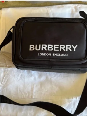 Svart axelväska från Burberry - Snygg svart axelväska från Burberry med tydlig logga och texten 'London England' på framsidan. Väskan har två stora fack med dragkedja och justerbar axelrem. Tillverkad i slitstarkt tygmaterial, perfekt för att bära det viktigaste.
