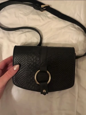 Svart midjeväska i läder - Snygg svart midjeväska i läder med ormmönstrad yta och guldfärgad ringdetalj framtill. Väskan har justerbar rem och stängs med magnetknapp. Perfekt för dig som vill ha en stilren men ändå trendig accessoar. Kolla min profil för fler plagg! 🥰