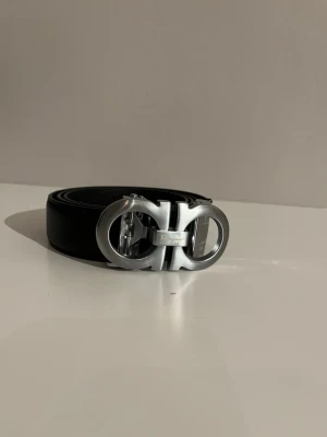 Svart skärp från Salvatore Ferragamo - Snyggt svart skärp från Salvatore Ferragamo med ikoniskt dubbel-Gancini-spänne i silverfärgad metall. Bältet är gjort i slätt skinn och har en stilren, modern look som passar till många outfits. Perfekt accessoar för dig som gillar exklusiva detaljer.