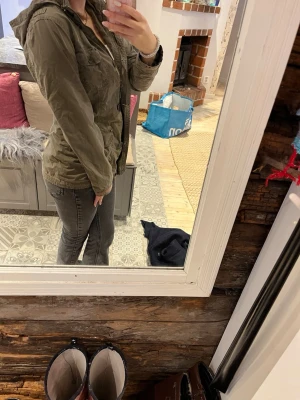 Olivgrön vindjacka med huva - Snygg olivgrön vindjacka med huva och dragkedja framtill. Jackan har flera fickor med lock och knappar, samt justerbara muddar vid ärmsluten. Perfekt för blåsiga dagar och enkel att matcha med olika outfits.