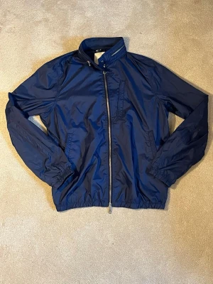 Marinblå vindjacka från J.Lindeberg - Snygg marinblå vindjacka från J.Lindeberg med dragkedja framtill, hög krage och elastiska muddar. Jackan har två sidofickor och är tillverkad i ett lätt, vindtätt material som passar perfekt för blåsiga dagar.