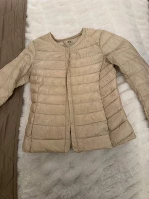 Beige lätt dunjacka från Uniqlo - Snygg beige dunjacka från Uniqlo med rund halsringning och tryckknappar framtill. Jackan är quiltad med horisontella sömmar och har en minimalistisk design utan huva. Perfekt för dig som gillar stilrena och lätta ytterplagg.