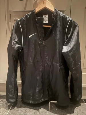 Svart Nike vindjacka XL junior  - Snygg svart vindjacka från Nike med vit logga på bröstet och tunna vita detaljer längs axlarna. Jackan har hel dragkedja framtill, ståkrage och meshfoder på insidan för extra komfort. Perfekt för blåsiga dagar och sportiga outfits.