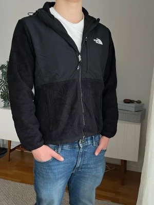 Svart The North Face Denali fleecejacka, storlek M - Svart The North Face denali fleecejacka med luva. Vit logga både bak och fram, justerbar luvstorlek och midja och många bra fickor.  Jackan är storlek L för damer men sitter perfekt som M på killar (se mått noga). Han på bild är 184 väger 80  Axelbredd: 46 cm Längd: 67 cm  Fleecematerialet: 100% polyester Skalmaterialet: 100% nylon  Mycket bra skick, inga defekter, jättefräsch 