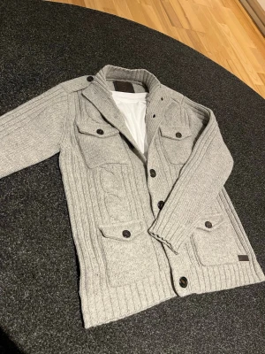 Grå stickad cardigan - Snygg grå stickad kofta med fyra stora fickor framtill och knappar längs hela framsidan. Koftan har ribbade muddar, hög krage och detaljerade sömmar. Perfekt för lager på lager och en chill look.