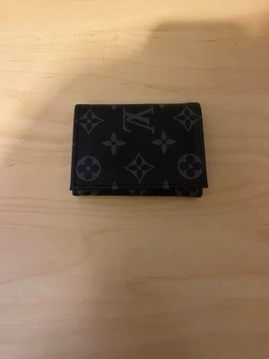 Monogram plånbol - Snygg svart plånbok  med klassiskt monogrammönster i grått. Tillverkad i tåligt läder och har en kompakt, vikbar design som gör den enkel att bära med sig. Perfekt för kort och sedlar.