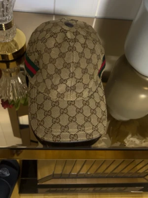 Gucci beige keps med GG-mönster - Säljer en beige keps från Gucci med det klassiska GG-monogrammet över hela kepsen. På sidan finns en grön och röd rand som ger en ikonisk touch. Kepsen har böjd skärm och är tillverkad i canvasmaterial. Perfekt för dig som vill ha en stilren och lyxig accessoar.