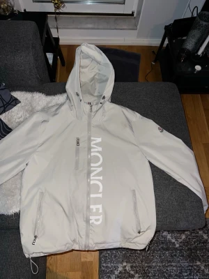 Moncler vindjacka - Snygg vit vindjacka från Moncler med stor logga tryckt vertikalt på framsidan. Jackan har huva, tre dragkedjefickor och justerbar nederkant. Perfekt för dig som vill ha en stilren och exklusiv look. Materialet är lätt och vattenavvisande. Den är knappt använd på grund av att den va för liten till mig. Perfekt för någon som är mellan 173-177cm lång