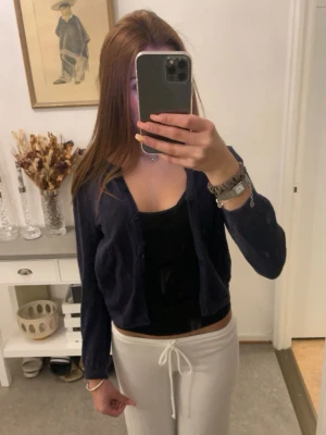 Mörkblå kofta med hålmönster - Snygg mörkblå kofta med hålmönster och knappar framtill. Kort modell med långa ärmar, perfekt att slänga över ett linne eller t-shirt. Liten rosa logga broderad på bröstet. Enkel och stilren design som passar till mycket.