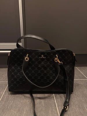 Svart handväska från Anna Field - Snygg svart handväska från Anna Field med präglat mönster och guldfärgade metalldetaljer. Väskan har både handtag och en justerbar axelrem, perfekt för att bära på olika sätt. Tillverkad i konstläder med stilren och modern design. Litet fack inuti. Mått: 27x38cm